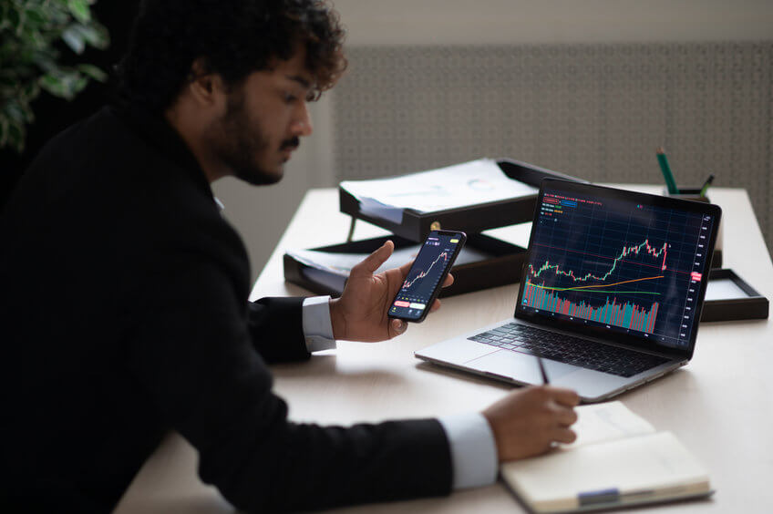 Smart focused Indian broker investor, stock trader using smartphone and laptop to analyze and invert in the financial market of cryptocurrencies, buy or sell crypto coins, planning strategy